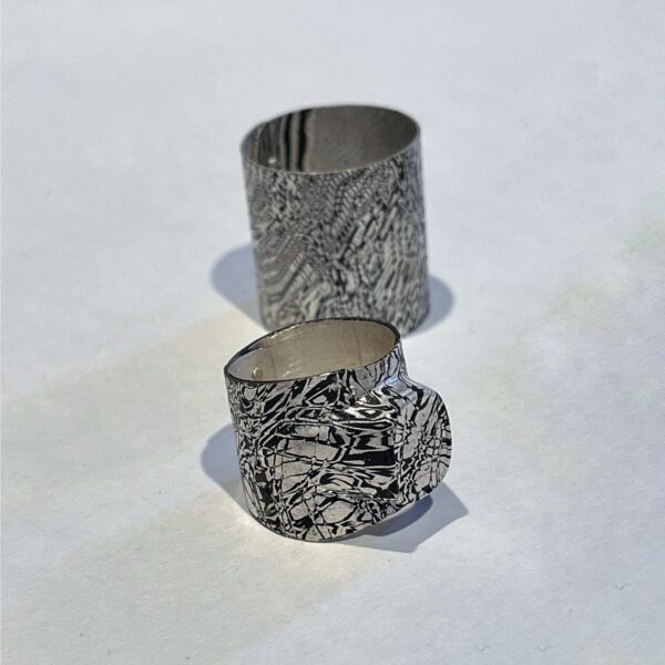 Zwei silbern-schwarze, strukturierte Metall-Ringe mit rauer, organisch anmutender Oberfläche, übereinander auf weißem Hintergrund fotografiert.