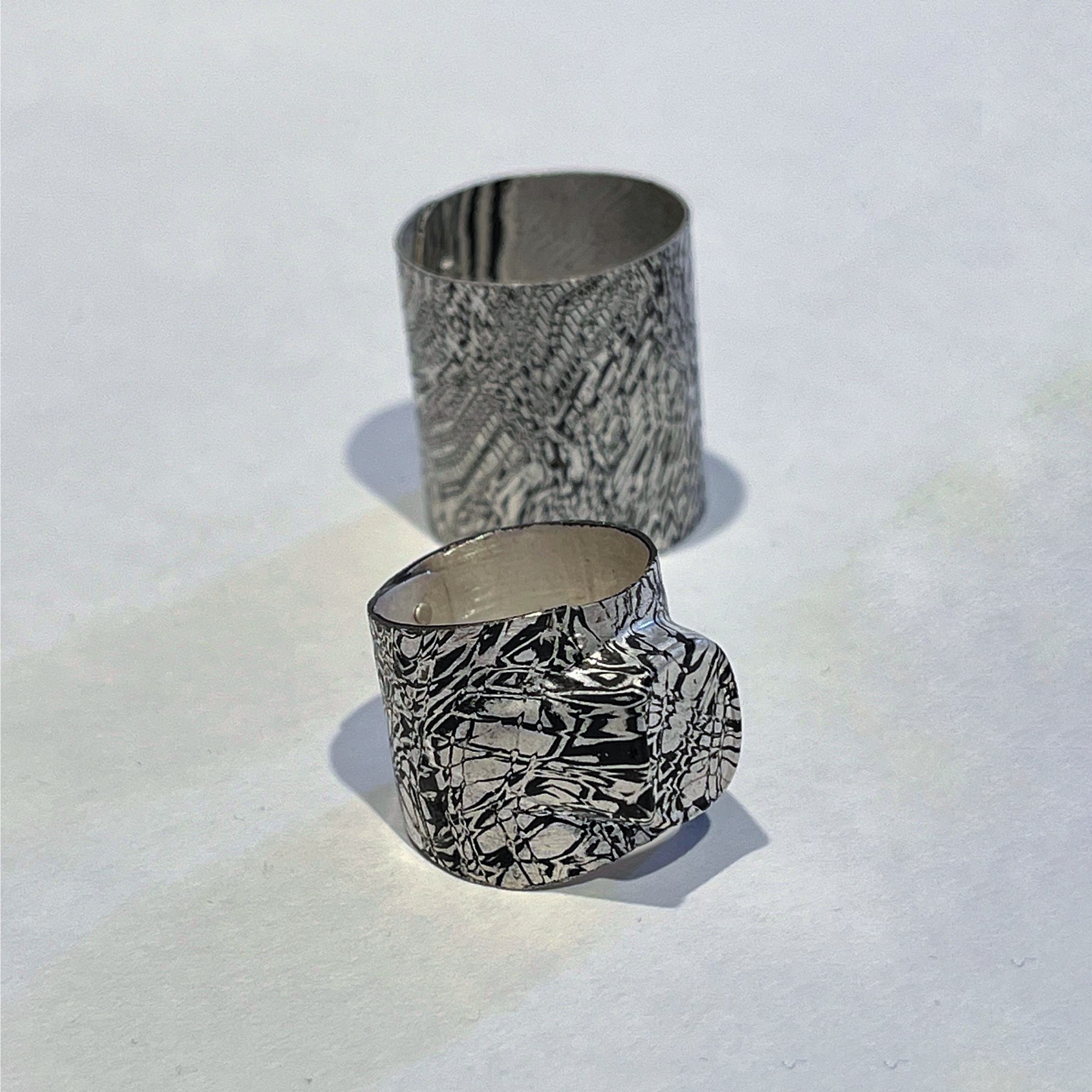 Zwei silbern-schwarze, strukturierte Metall-Ringe mit rauer, organisch anmutender Oberfläche, übereinander auf weißem Hintergrund fotografiert.