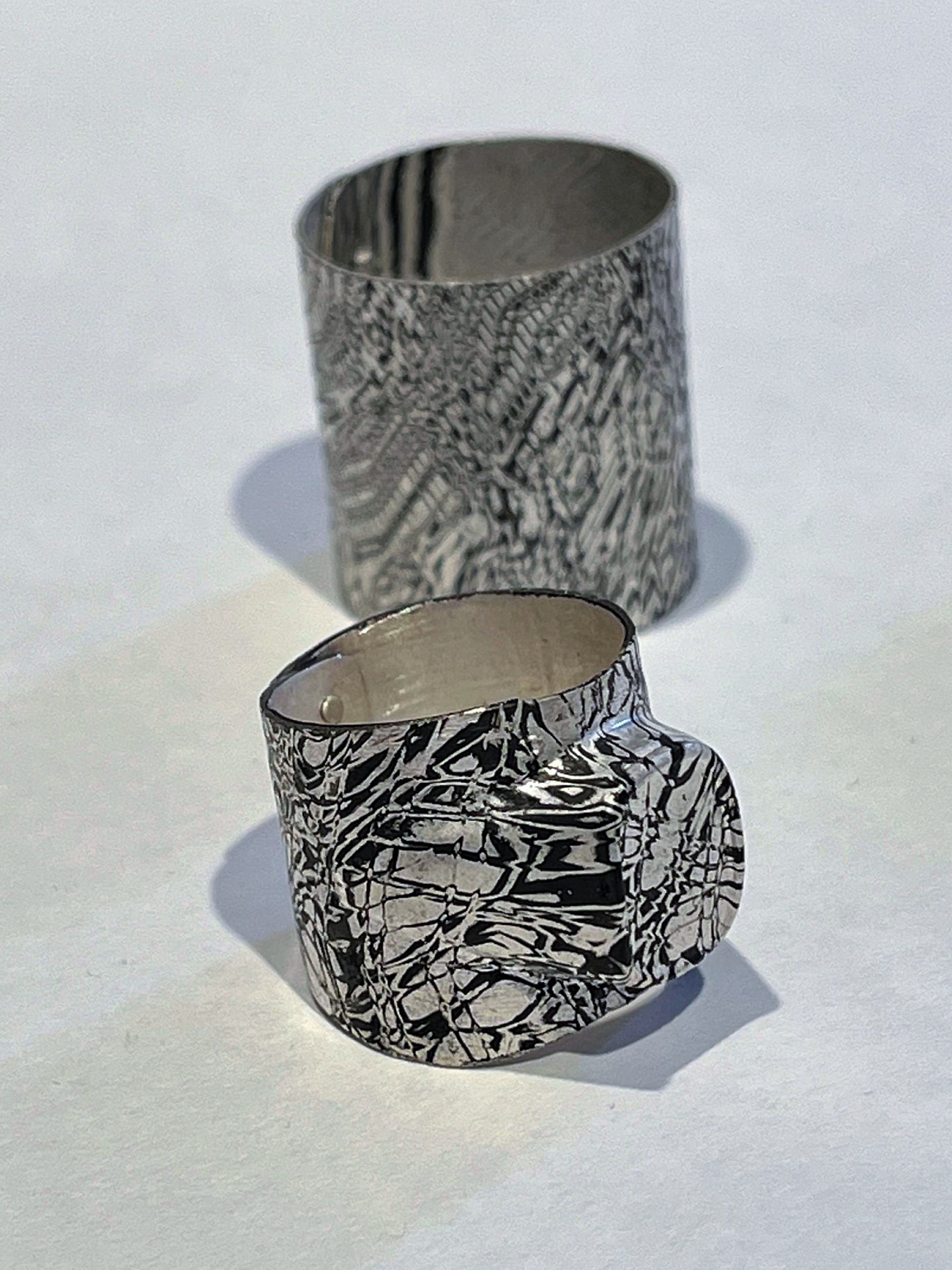 Zwei silbern-schwarze, strukturierte Metall-Ringe mit rauer, organisch anmutender Oberfläche, übereinander auf weißem Hintergrund fotografiert.
