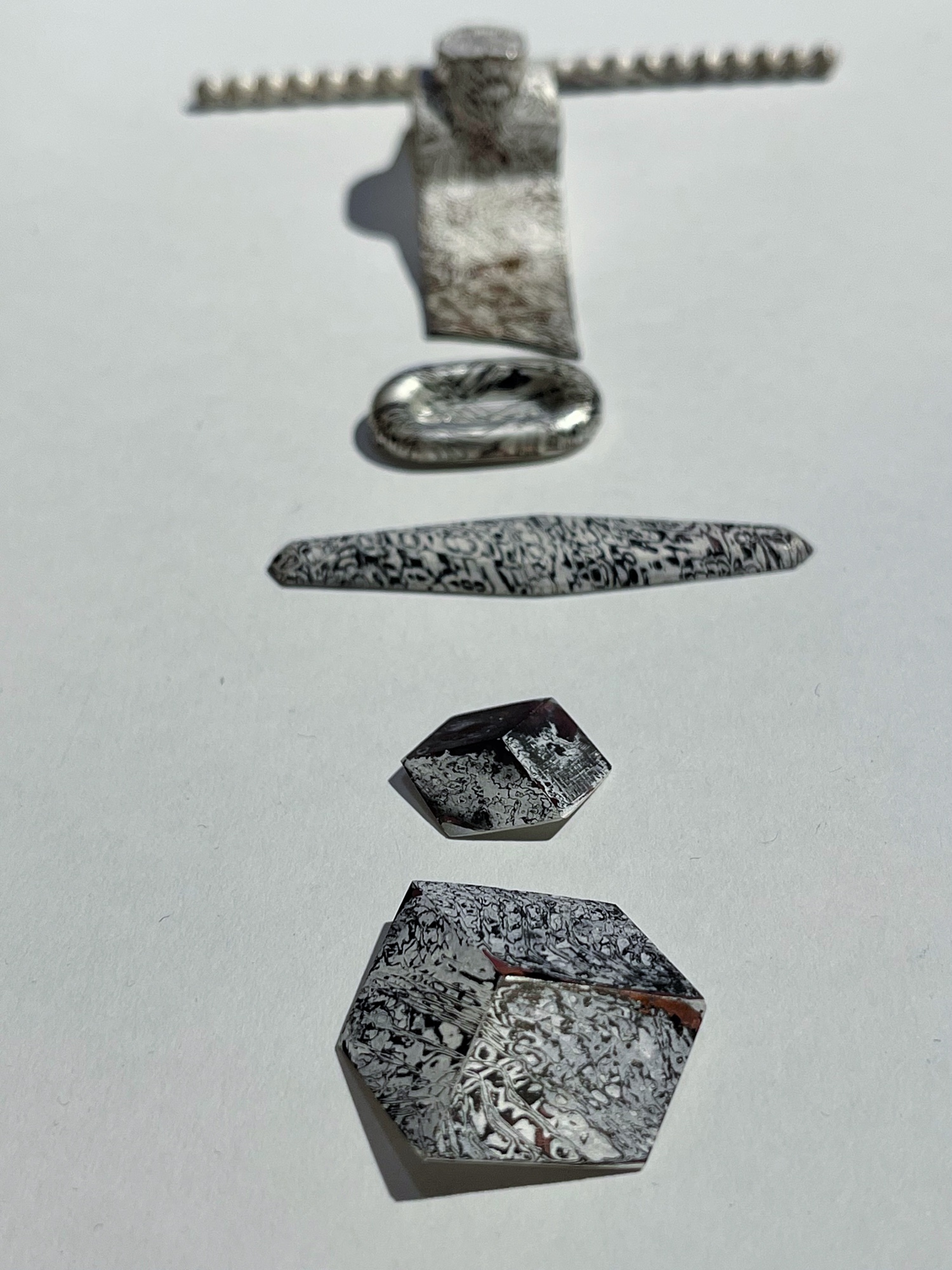 Sechs broschenartige Schmuckstücke mit silber-schwarz strukturierter Oberfläche, die organisch und rau anmutet.
