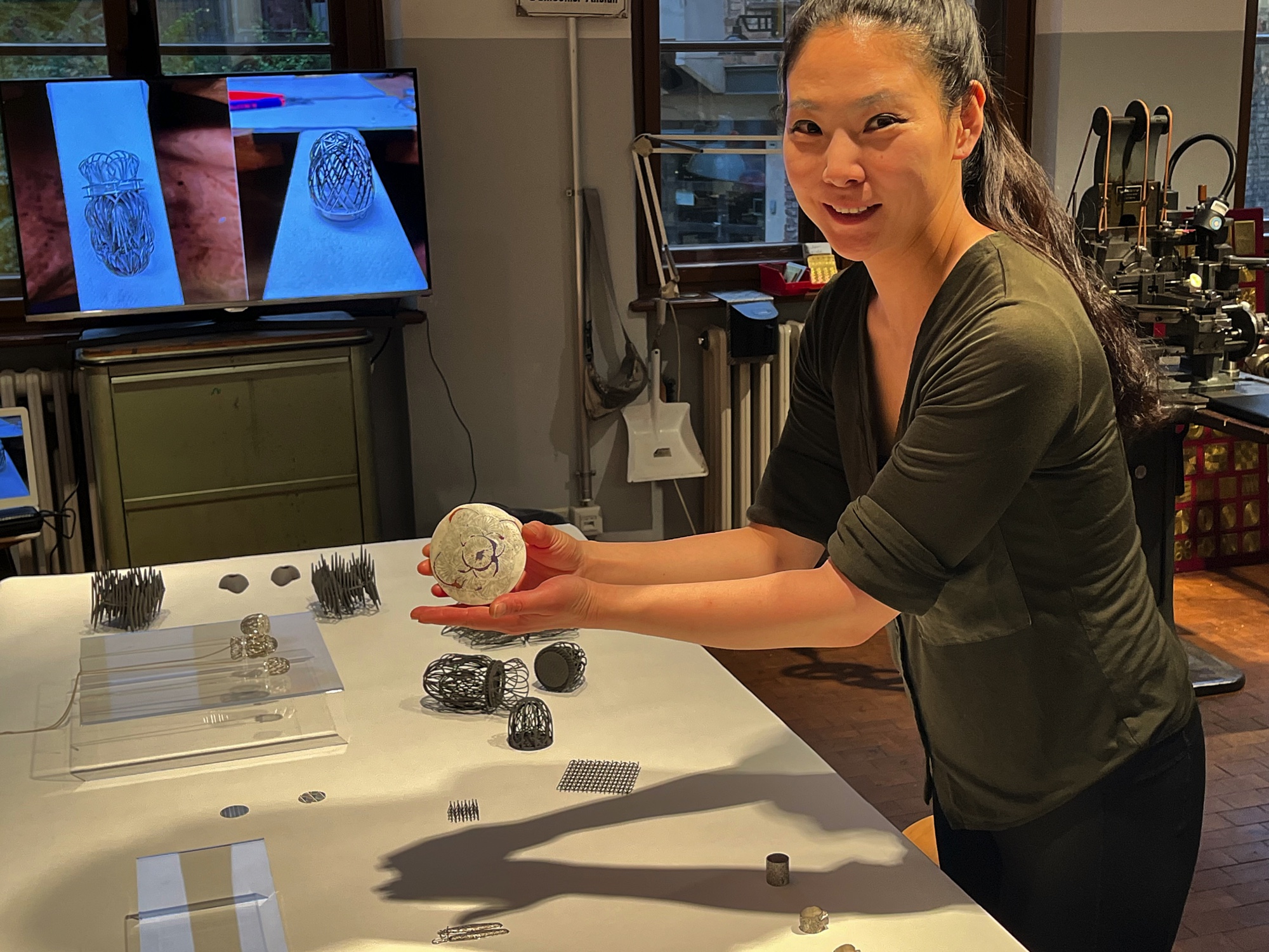 Lächelnde Frau in einer Werkstatt hält einen abgerundeten, steinartigen Gegenstand, im Hintergrund 3D-Druck-Modelle und zwei Bildschirme mit Schmuckstücken.