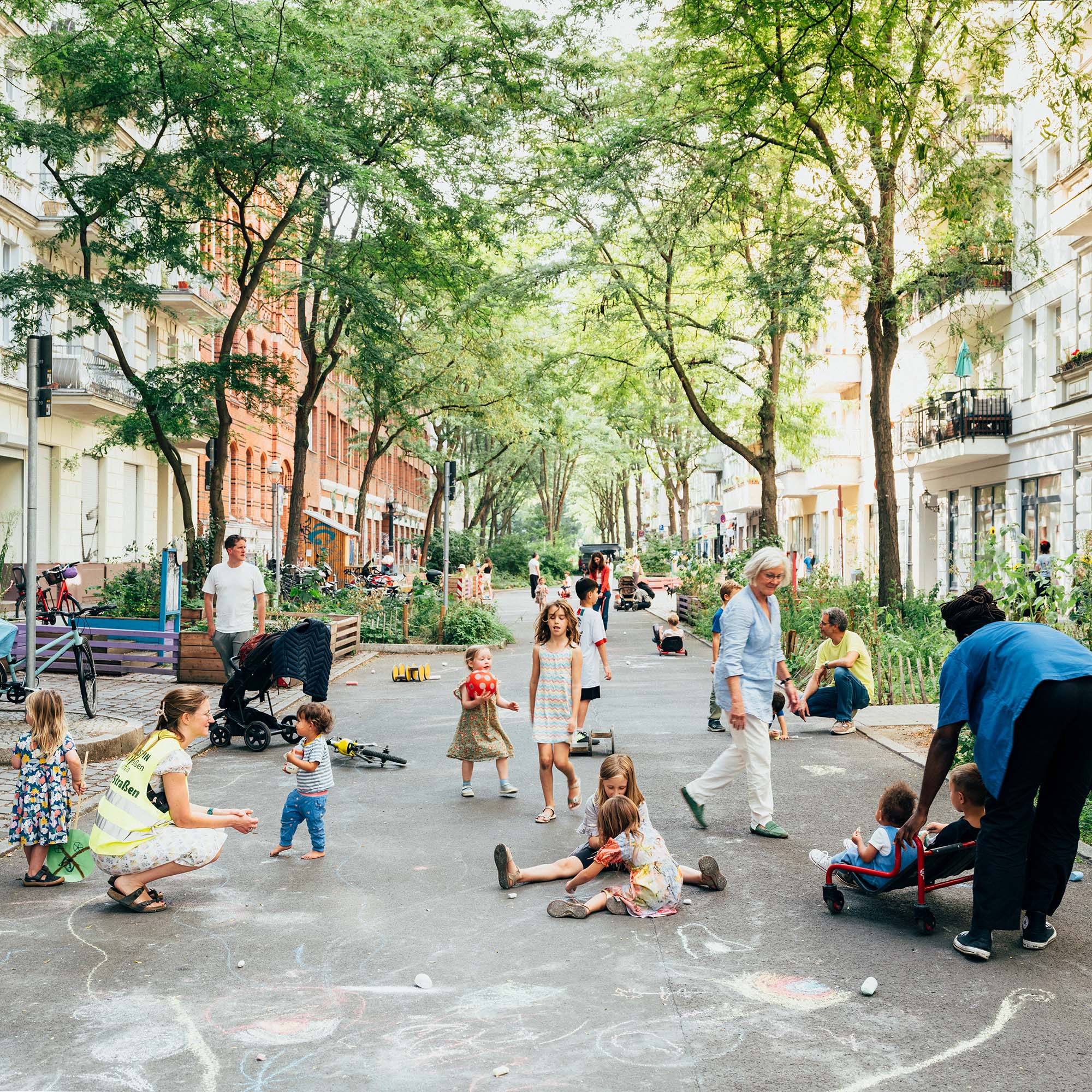 Belebte Straße mit spielenden Kindern, Erwachsenen, Kinderwagen, Fahrrädern und Bäumen in einer urbanen Umgebung.