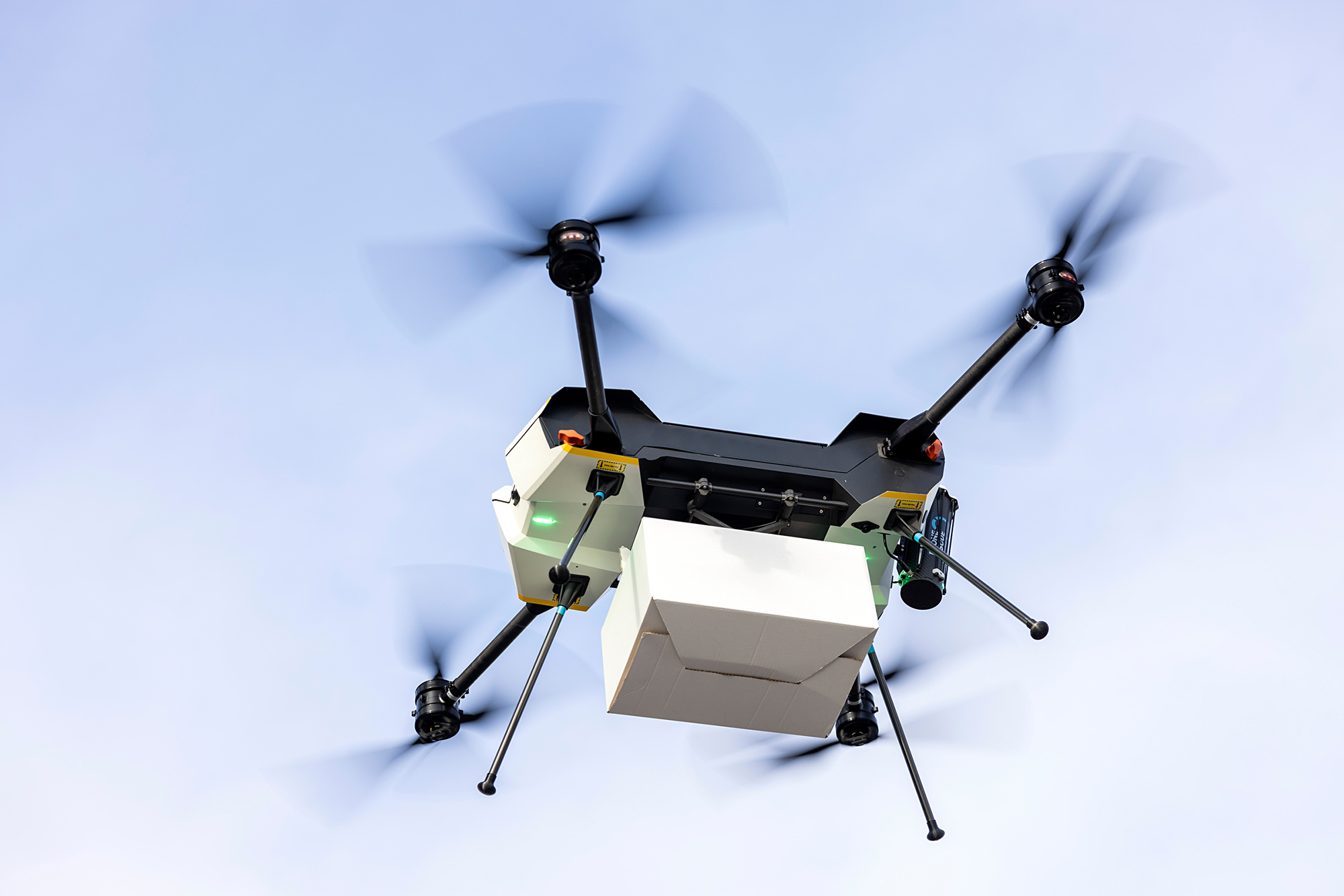 Drohne mit vier Propellern, die ein weißes Paket transportiert.