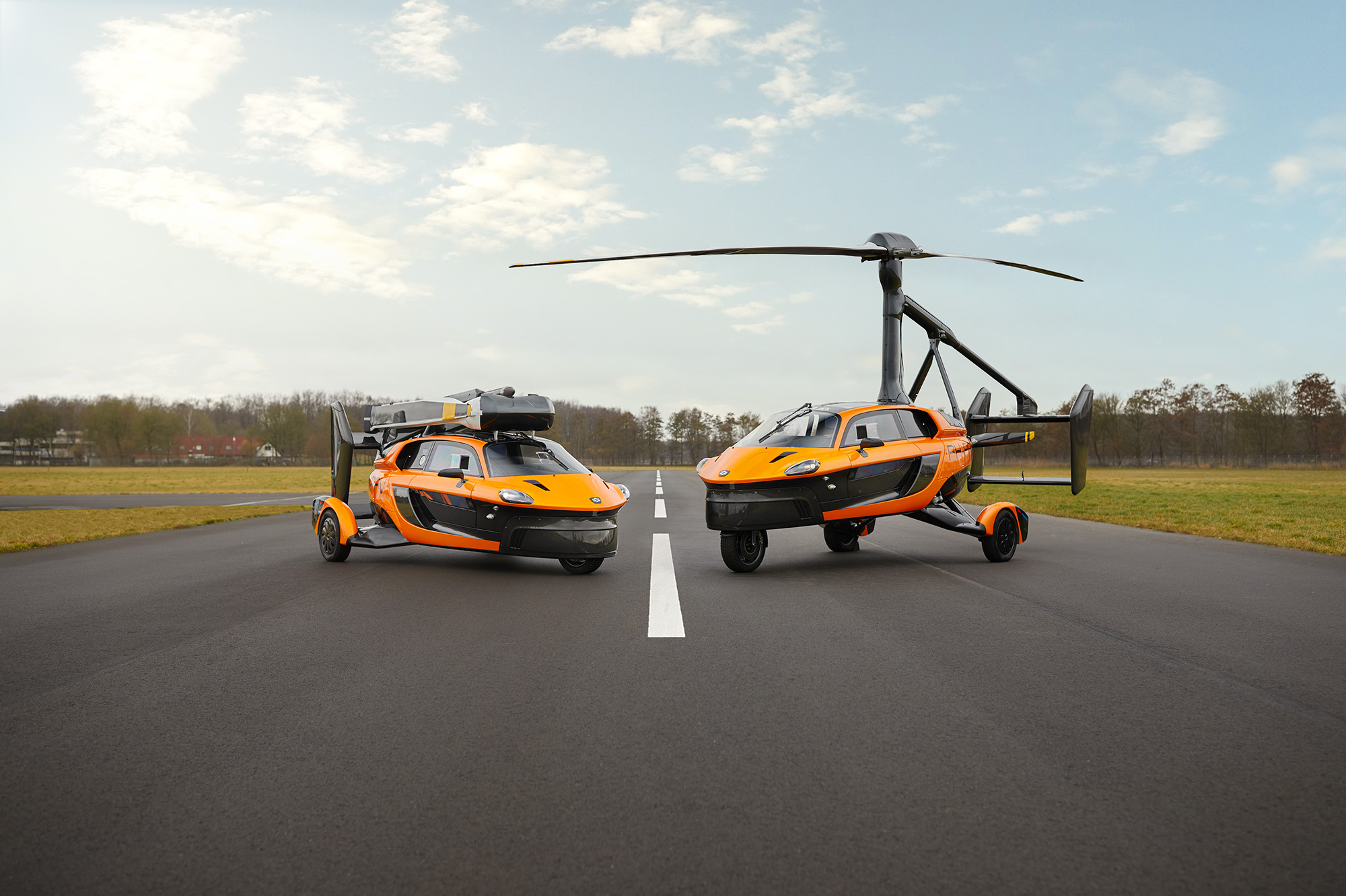 Zwei orange-schwarze Flugautos mit Propellern auf einer Landebahn vor einer grünen Landschaft.