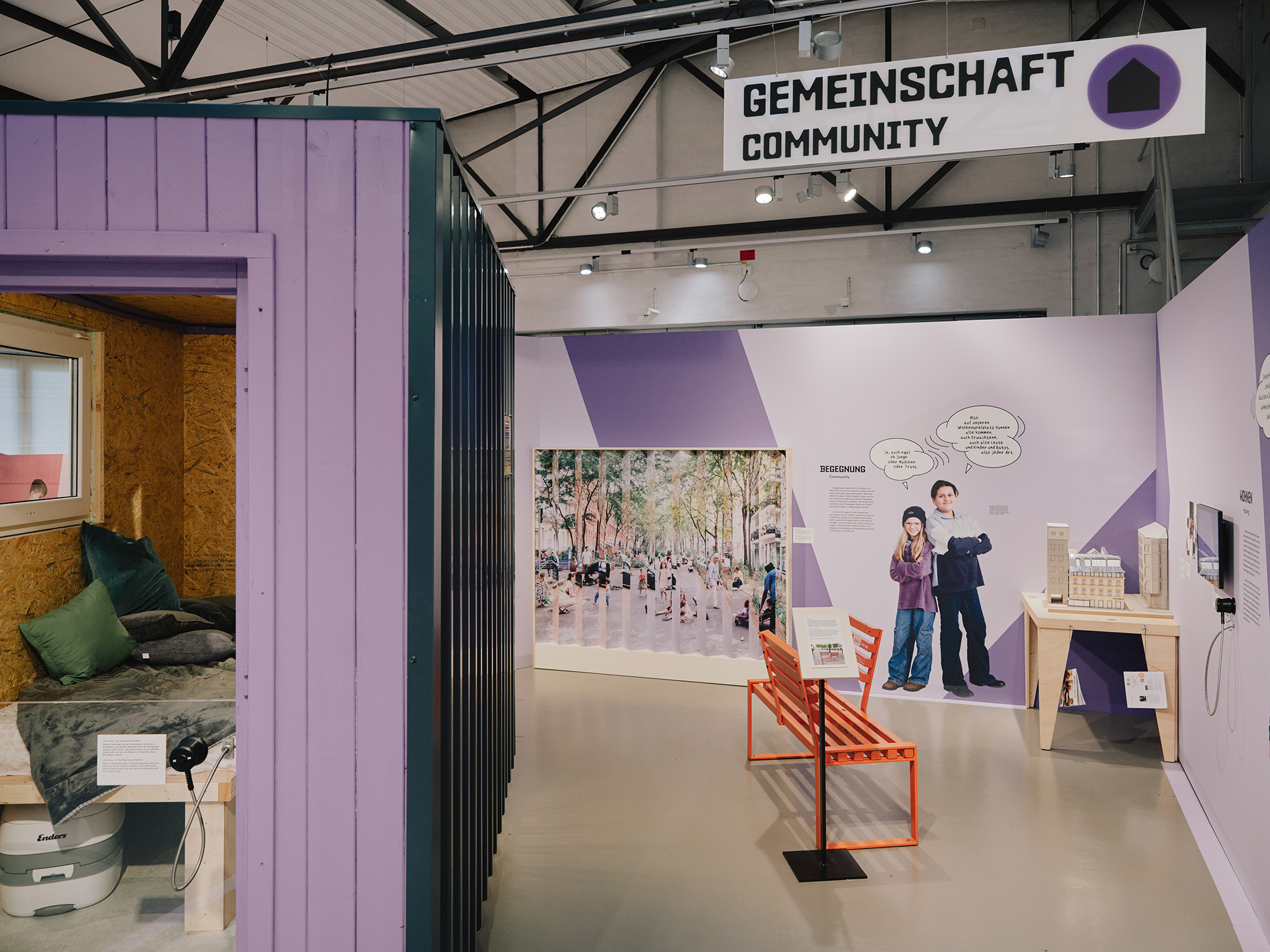Ausstellungsbereich mit dem Titel "Gemeinschaft Community", der einen Wohncontainer mit Bett und Kissen, eine orange Bank, Infotafeln und ein großes Foto mit zwei Personen zeigt.