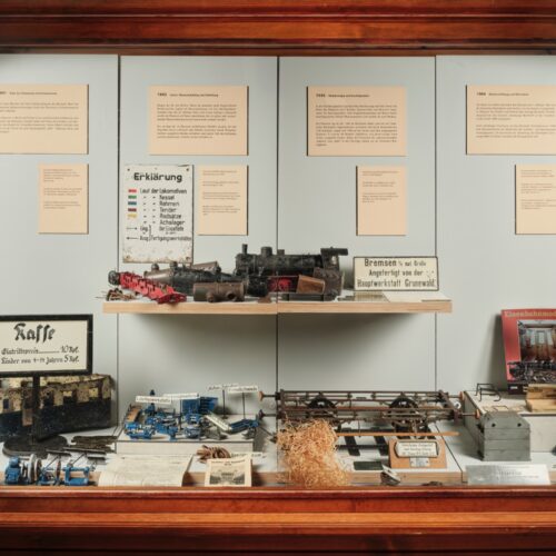 Vitrine mit historischen Eisenbahnmodellen, Lokomotivteilen, Werkzeugen und Infotafeln zur Geschichte einer Werkstatt; Exponate sind geordnet auf mehreren Ebenen präsentiert.