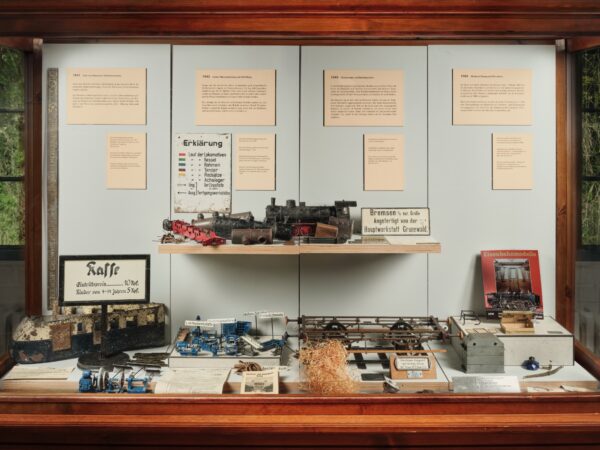 Vitrine mit historischen Eisenbahnmodellen, Lokomotivteilen, Werkzeugen und Infotafeln zur Geschichte einer Werkstatt; Exponate sind geordnet auf mehreren Ebenen präsentiert.