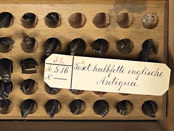 Holzschublade mit vielen runden Vertiefungen, gefüllt mit alten Druckstempeln, beschriftet: "Text halbfette englische Antiqua."