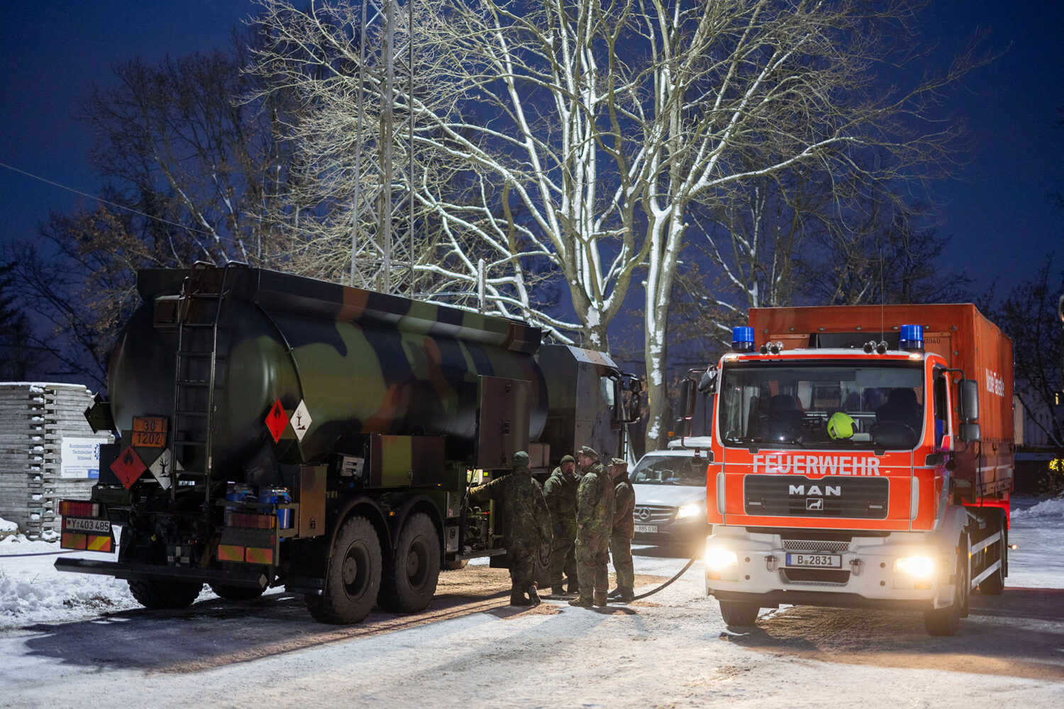 Nachtaufnahme: Ein Militär-Tankwagen und ein rotes Feuerwehrfahrzeug der Marke MAN mit Blaulicht stehen auf einer verschneiten Straße. Im Hintergrund sind Soldaten und kahle Bäume zu sehen.