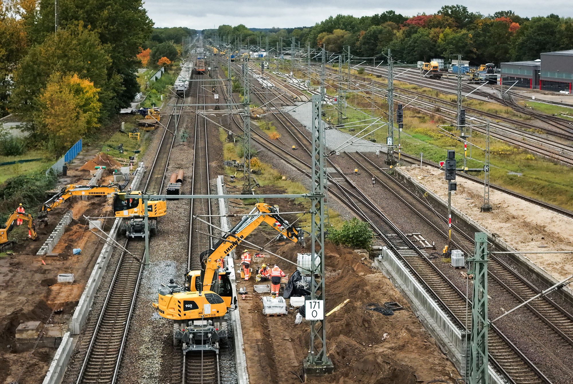 Bauarbeiten an einem Bahnhofsgleis: Gelbe Baumaschinen von Liebherr und Arbeiter in orangefarbener Schutzkleidung arbeiten an den Schienen, während im Hintergrund weitere Gleise und ein Wald zu sehen sind.