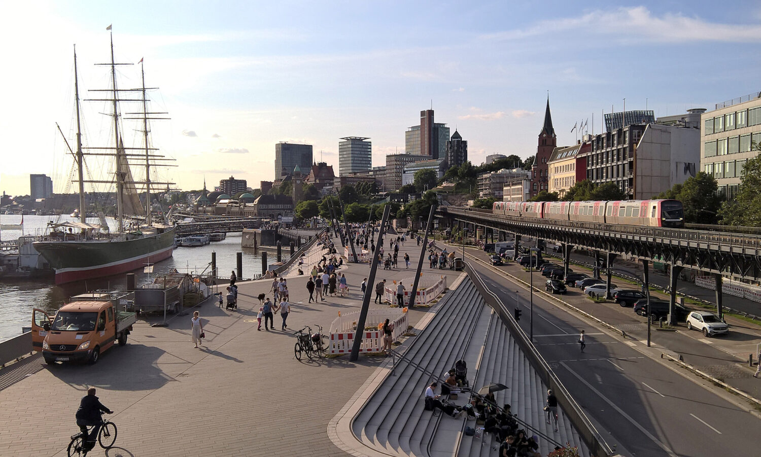 Stadtpanorama mit einem Segelschiff, einer belebten Promenade, einer U-Bahn auf einer Brücke, modernen Gebäuden, einer Kirche mit spitzem Turm und einem orangefarbenen Lieferwagen im Vordergrund. Im Hintergrund sind Hochhäuser und der Hafen zu sehen.