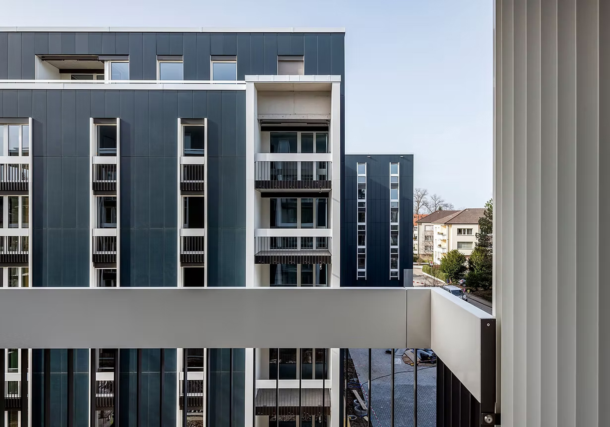 Moderne Wohngebäude mit dunkler Fassade, weißen Fensterrahmen und Balkonen. Im Hintergrund sind ältere Häuser mit schrägen Dächern zu sehen.