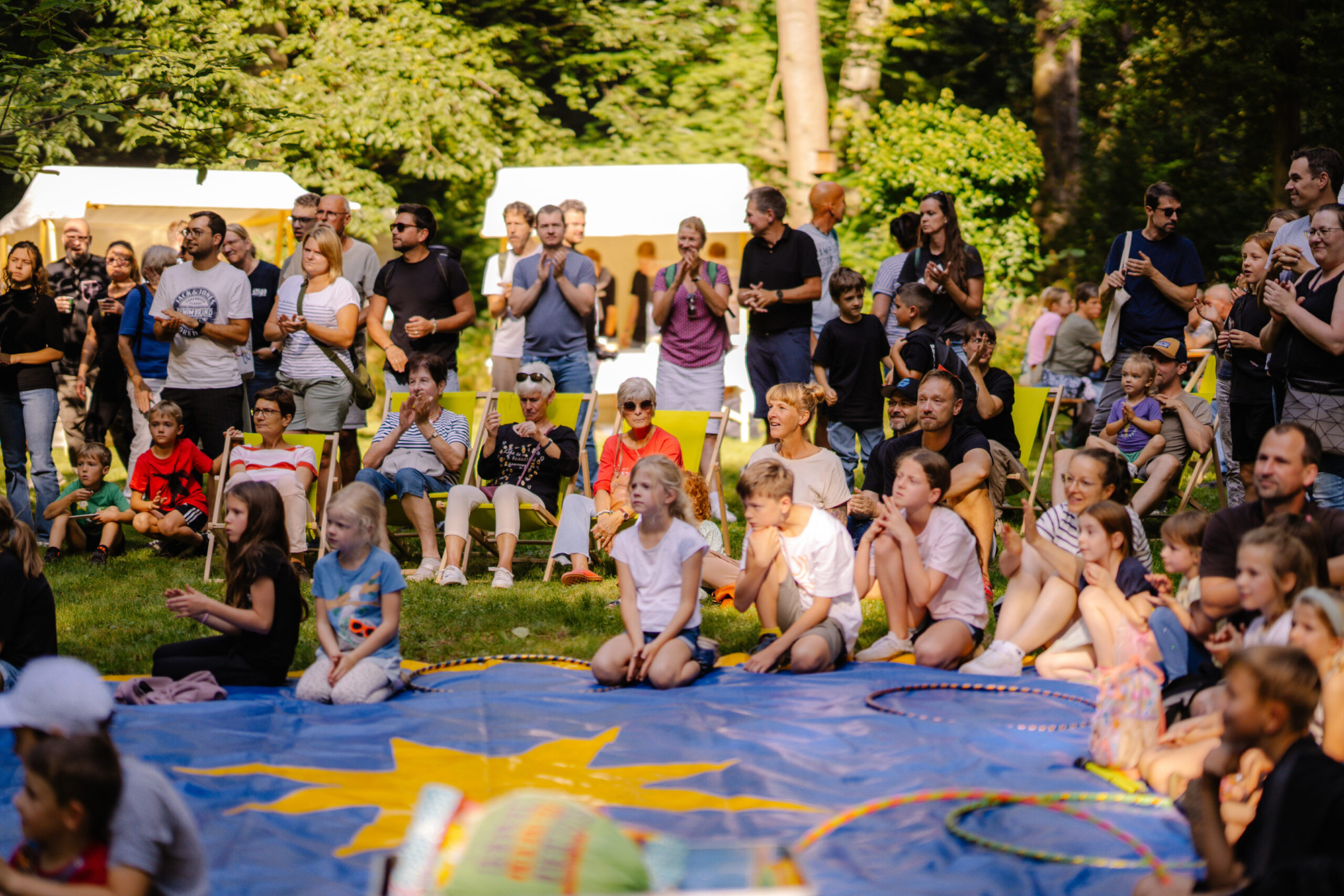 Gruppe von Menschen, die auf Klappstühlen und Decken im Freien sitzen und einer Vorstellung auf einer blauen Plane zuschauen.
