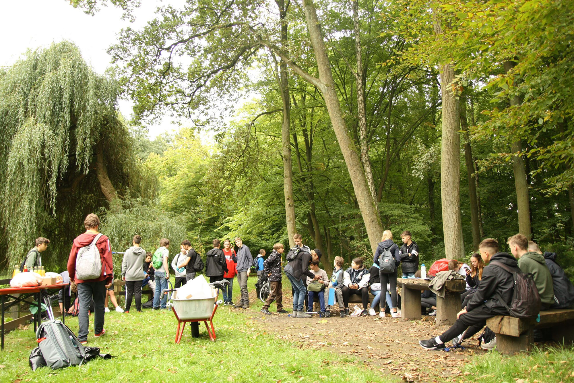 Gruppe von Jugendlichen und jungen Erwachsenen in einem Wald mit Tischen, Bänken, Rucksäcken und einer Schubkarre.