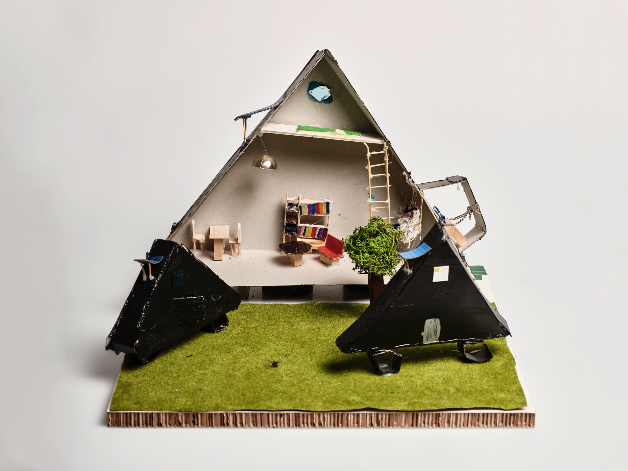 Modell eines dreieckigen Hauses mit abnehmbaren Dachteilen, Möbeln, einem Baum und grünem Boden auf einer Pappbasis.