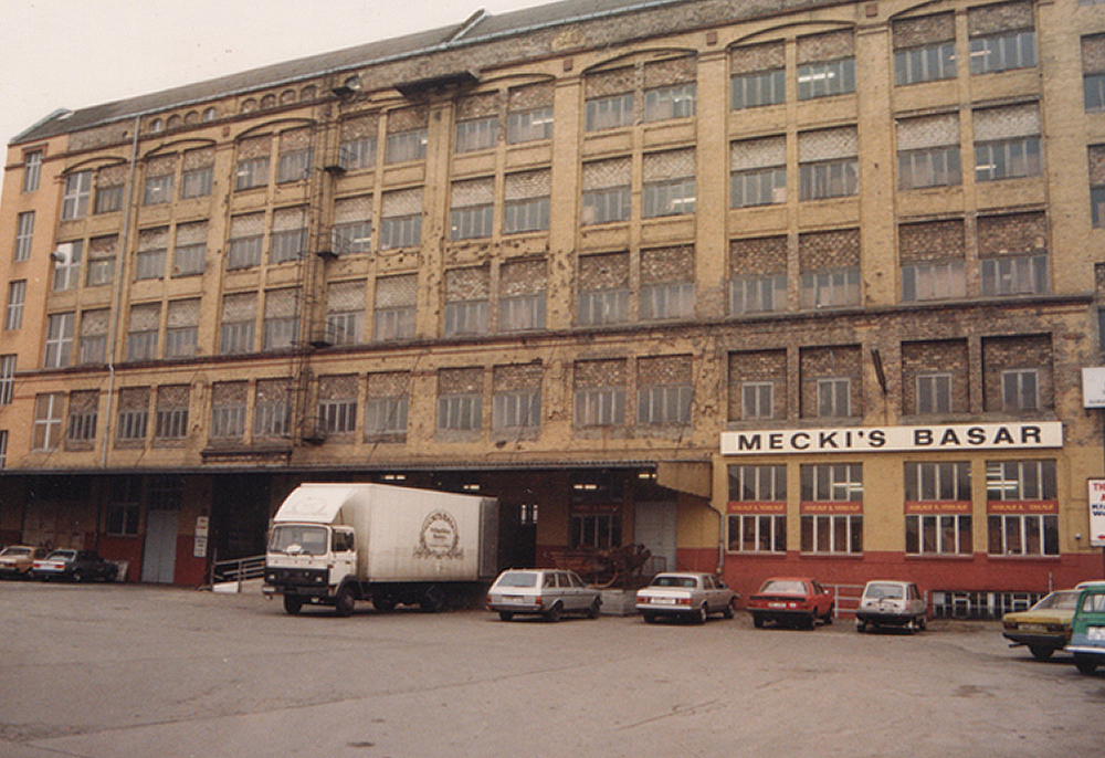 Historisches Fabrikgebäude mit der Aufschrift 'Mecki’s Basar', davor ein LKW und geparkte Autos.