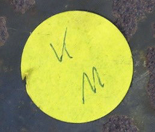 Gelber runde Aufkleber mit handschriftlichem ‚V‘ und ‚M‘ auf dunklem Untergrund.