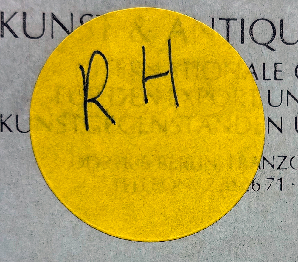 Nahaufnahme eines gelben, runden Aufklebers mit der handschriftlichen Aufschrift „RH“ auf einem gedruckten Dokument.