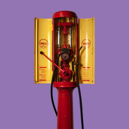 Historische rote und gelbe Shell-Benzinzapfsäule mit sichtbarer Mechanik, Schläuchen und Messanzeige vor violettem Hintergrund.