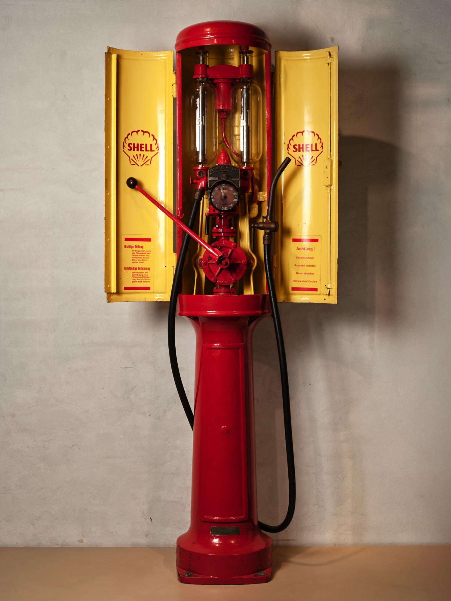 Historische rote Shell-Zapfsäule mit gelben Flügeltüren, sichtbaren Pumpen, Schläuchen und Messanzeige vor einer Wand.