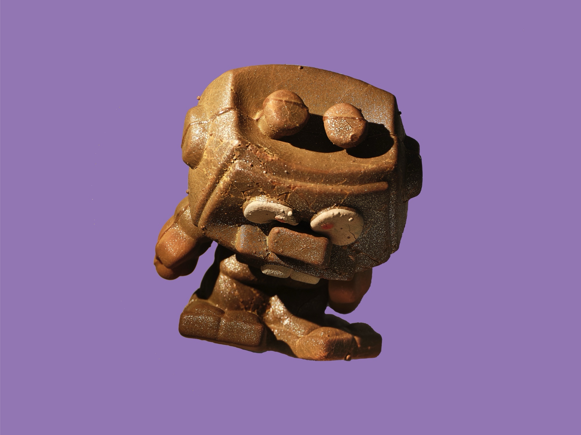 Eine skulpturhafte Figur aus einem glänzenden, goldbraunen Material: ein kubischer Körper mit zwei hervorstehenden Augen und einem schlitzförmigen Mund, dazu würfelförmige Arme und Beine, vor lilafarbenem Hintergrund.